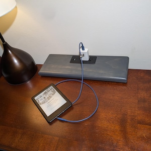 Puede incluir: Una mesita de noche de madera marrón oscuro con una lámpara y un lector electrónico. Una estación de carga de madera gris con un cargador blanco está enchufada a la pared. El lector electrónico está conectado al cargador con un cable azul.