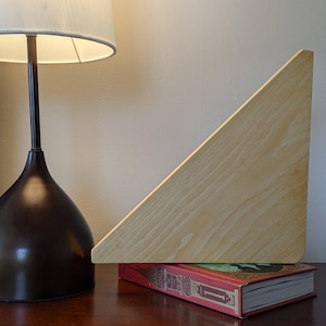 Puede incluir: Un separador de libros triangular de madera clara con bordes redondeados, que sostiene un libro rojo con letras doradas sobre una superficie de madera.