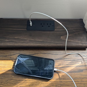 May include: A dark stained wooden charging station with a black power outlet and a smartphone charging. The phone is black with a black case. A white charging cable is plugged into the phone and the outlet.