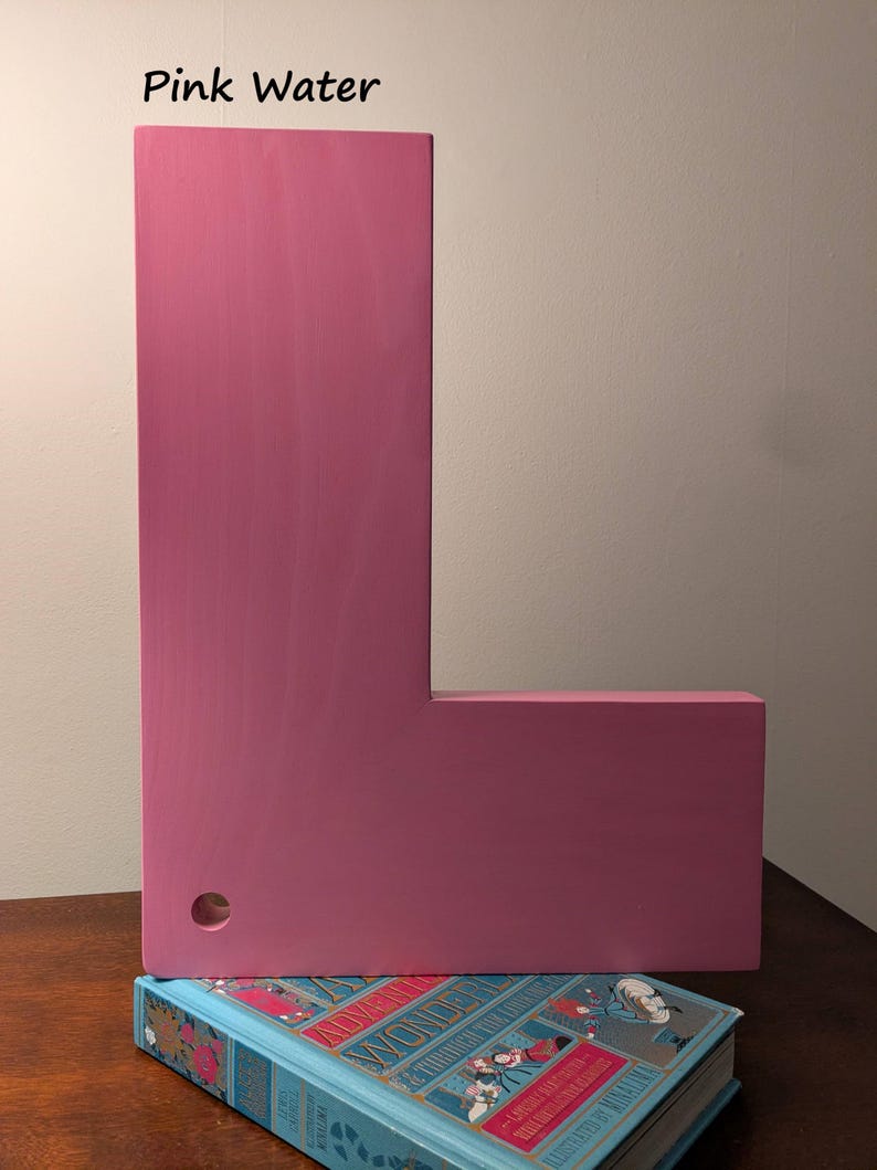 May include: A large, pink wooden letter "L" stands upright, with a small hole drilled into the side. The letter rests on a blue book with gold lettering. The text "Pink Water" is above the letter.