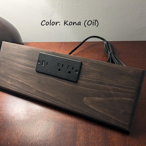 May include: A rectangular, stained wooden power strip with a black outlet and USB port panel. The wood is a dark brown colour, and the power strip has a black cord. The text "Color: Kona (Oil)" is visible.