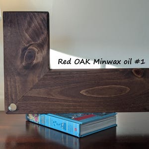 May include: A dark brown, L-shaped wooden shelf, stained with Red Oak Minwax oil #1, rests on a blue book. The wood grain is visible. A silver-colored metal disc is visible on the corner of the shelf.