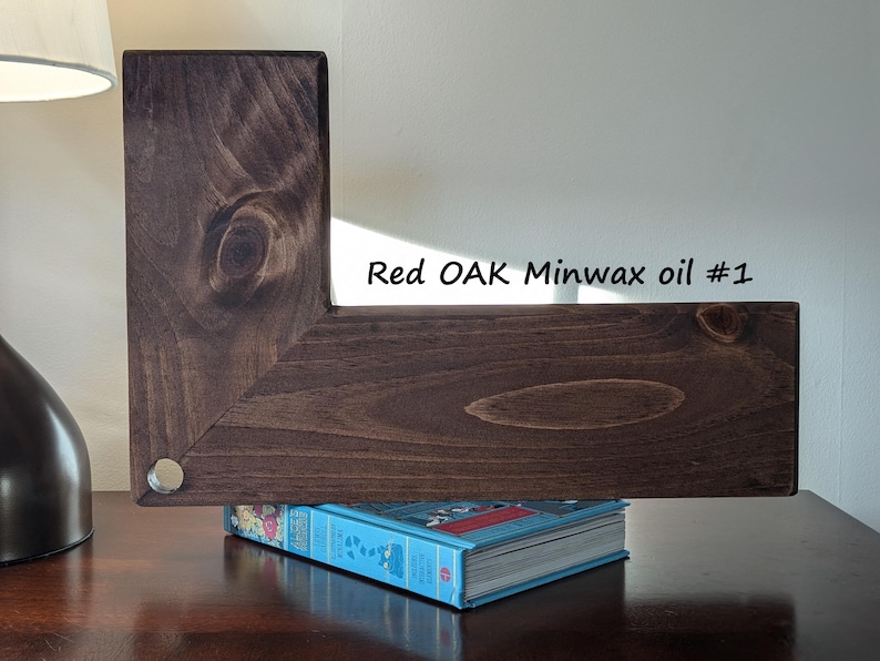 May include: A dark brown, L-shaped wooden shelf, stained with Red Oak Minwax oil #1, rests on a blue book. The wood grain is visible. A silver-colored metal disc is visible on the corner of the shelf.