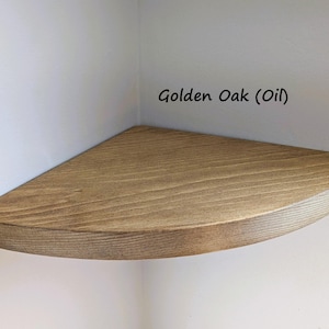 May include: A corner shelf made of golden oak wood, installed in a white corner. The shelf has a rounded front edge and is labeled "Golden Oak (Oil)". The wood grain is visible.