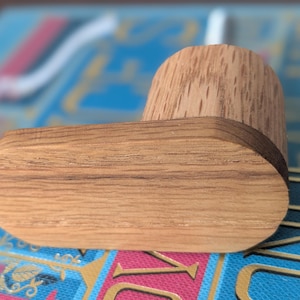 Puede incluir: Una pipa de madera con un cuenco redondeado y una base rectangular plana. La pipa está hecha de madera de color marrón claro y tiene un acabado liso. La pipa descansa sobre una superficie estampada en azul y dorado.