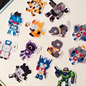 May include: A collection of colorful cartoon stickers featuring various Transformers characters. The stickers showcase characters in different poses and colors, including blue, orange, gray, and green. The stickers are on a white background.