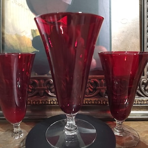 Puede incluir: Tres copas de vino de color rojo rubí con un diseño acampanado. Las copas tienen una base clara y circular y un tallo detallado. El fondo incluye una imagen enmarcada con un borde dorado y una superficie negra.