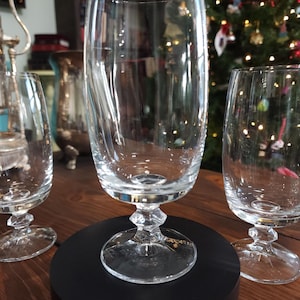 May include: Clear glass stemware with a diamond-shaped base and a tall, slender bowl. The glass is displayed on a black circular stand, with other glasses visible in the background. The stemware is likely for serving beverages.