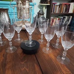 Puede incluir: Un conjunto de copas de vino de cristal transparente con un diseño texturizado en el cuenco y un tallo de cuentas. Las copas están dispuestas sobre una mesa de madera oscura, con una jarra y una cubitera plateadas al fondo.