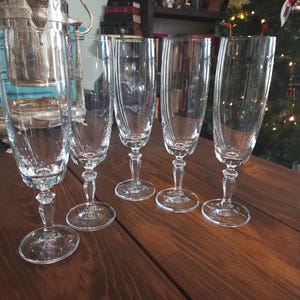 May include: Five clear champagne flutes with decorative stems and bases are arranged on a dark wooden surface. The flutes have a tall, slender design with vertical etched lines. A silver ice bucket and a Christmas tree are in the background.
