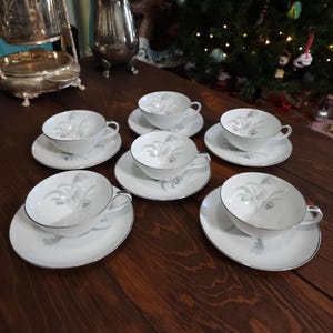 May include: A set of six white teacups and saucers with a silver floral design and silver trim. The teacups and saucers are arranged on a dark wooden surface. The teacups have handles.