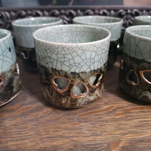 May include: A set of ceramic cups featuring a pale green crackle glaze on the upper portion and a dark, textured brown base. Each cup is adorned with a series of small, triangular cutouts with metallic gold-toned edges around the lower section.