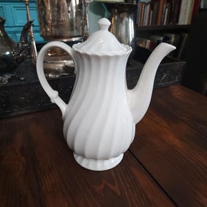 Op de afbeelding: Witte keramische theepot met een spiraalvormig ontwerp, een gebogen handvat en een tuit. De theepot heeft een deksel met een kleine knop bovenop. De theepot staat op een donkere houten ondergrond.