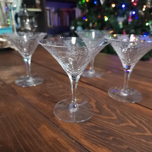 May include: Four clear, etched glass martini glasses with long stems sit on a dark wooden surface. The glasses have a classic triangular bowl shape and are decorated with a delicate floral pattern. The background includes a Christmas tree with colorful lights.