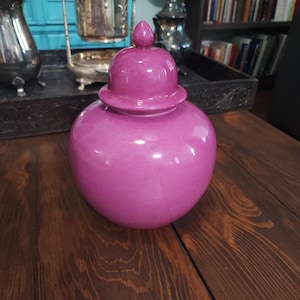 May include: A magenta-colored ceramic ginger jar with a matching lid. The jar has a rounded body and a slightly flared base. The lid is topped with a small, rounded finial. The jar is sitting on a dark wooden surface.