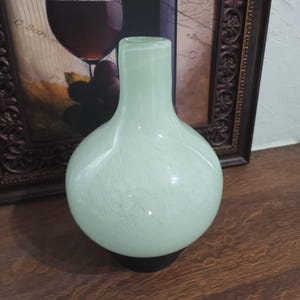 May include: A pale green glass vase with a rounded body and a cylindrical neck. The vase has a speckled pattern and sits on a black base. The background includes a framed wine-themed print.