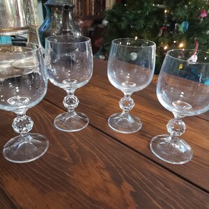 May include: Four clear wine glasses with a faceted crystal-like design on the stem, sitting on a dark wooden surface. The glasses have a rounded bowl and a clear, circular base. A Christmas tree is visible in the background.