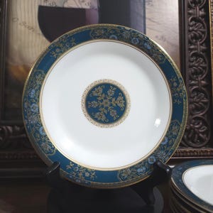 Puede incluir: Un plato blanco con un borde floral turquesa y dorado y un diseño central. El plato se exhibe sobre un soporte negro, con una pila de platos similares visibles en el fondo. El plato tiene un borde dorado.