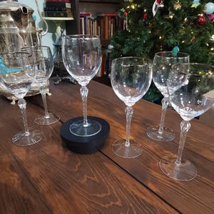 May include: A collection of clear crystal wine glasses with twisted stems, arranged on a dark wooden table. The glasses are empty and reflect the light. A silver tea set and a Christmas tree are in the background.