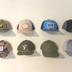 May include: Eight different baseball caps with various designs and colors. The caps are hanging on a wall. The designs include "Y'all", "Southern","Hooey", "Duck Commander", "HD", "Local", and "All Horse".