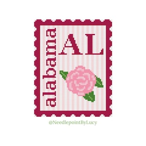 May include: A cross-stitch design in the shape of a postage stamp, with the word "Alabama" and the abbreviation "AL" in burgundy. A pink rose with green leaves is in the center. The background has pink and white stripes. The design is framed by a burgundy border.