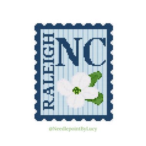 May include: A cross-stitch design of a postage stamp featuring "NC" in navy blue, with "RALEIGH" vertically on the left. A white dogwood flower with green leaves is in the center. The background is light blue.