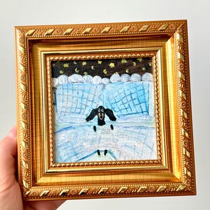 Puede incluir: Pintura enmarcada que representa una oveja en blanco y negro en una cama con ropa de cama azul. El marco dorado tiene un borde detallado. El fondo de la pintura representa un cielo nocturno con estrellas.