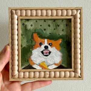 May include: A framed painting of a smiling corgi dog with orange and white fur, holding a piece of food. The artwork is set against a green background and framed in a light wood frame with a beaded edge.