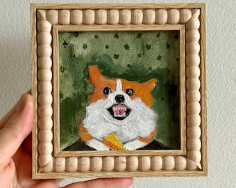 May include: A framed painting of a smiling corgi dog with orange and white fur, holding a piece of food. The artwork is set against a green background and framed in a light wood frame with a beaded edge.