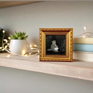 May include: A small, square, gold-framed painting of a cat. The cat is gray and white, holding a white cloth. The painting is displayed on a wooden shelf with a succulent plant in a white pot and a string of lights.