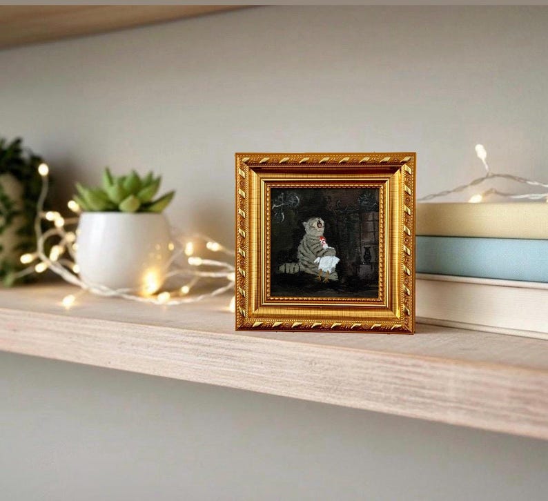 May include: A small, square, gold-framed painting of a cat. The cat is gray and white, holding a white cloth. The painting is displayed on a wooden shelf with a succulent plant in a white pot and a string of lights.