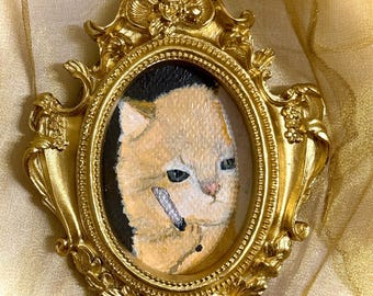 May include: A gold-colored, ornate picture frame with an oval-shaped painting of a cat. The cat is light orange with blue eyes and a black and white object in its mouth. The frame has decorative flourishes.