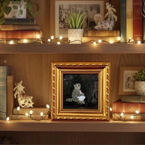 May include: A framed painting with a gold ornate frame. The painting depicts a gray tabby cat holding a white cloth. The painting is displayed on a wooden shelf with books, plants, and decorative lights.