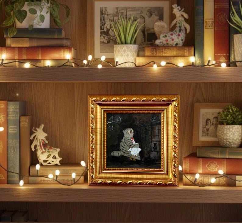 May include: A framed painting with a gold ornate frame. The painting depicts a gray tabby cat holding a white cloth. The painting is displayed on a wooden shelf with books, plants, and decorative lights.