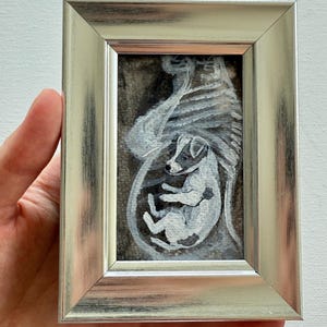 May include: A small framed painting featuring a white and gray dog inside a rib cage. The artwork is set against a dark background and framed in a silver-colored frame. The dog is painted in a realistic style.