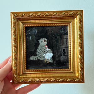 May include: A framed painting depicts a gray tabby cat holding a white chicken. The cat is in a dark room with a fireplace. The painting is in a gold frame with a detailed border.