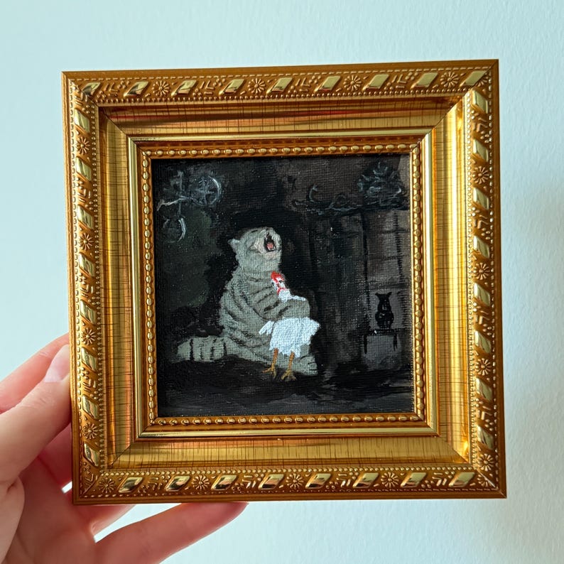 May include: A framed painting depicts a gray tabby cat holding a white chicken. The cat is in a dark room with a fireplace. The painting is in a gold frame with a detailed border.