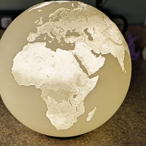 May include: A glowing globe lamp with a detailed relief map of the world. The continents are highlighted in a lighter color, creating a striking contrast against the globe's overall hue. This decorative lamp is a unique home decor item.
