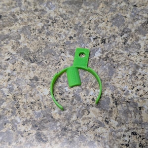 May include: A bright green plastic clip with a curved design. The clip has a rectangular top with a hole and a cylindrical piece in the middle. The clip is designed to hold something.