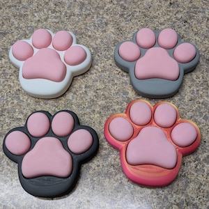 May include: Four paw-shaped silicone fidget toys in different colors: white, gray, black, and orange. Each toy has five pink dimples.