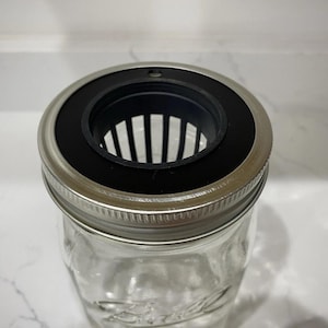 May include: A clear glass mason jar with a silver metal lid and a black plastic insert. The insert has vertical slots. The jar is designed for home and kitchen use, possibly for food storage or canning.