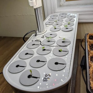 May include: White hydroponic gardening system with 16 individual plant pods. The system has a white metal pole with a clip on the top. The system has a black power cord. The system has the letters "RS" written on the side. There are several small green plants growing in the pods.