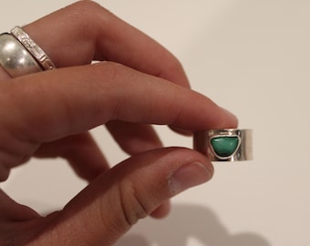 May include: A silver ring with a turquoise stone. The ring has a wide band and a bezel setting that holds the polished, green-blue stone. Another silver ring is visible on the finger in the background.