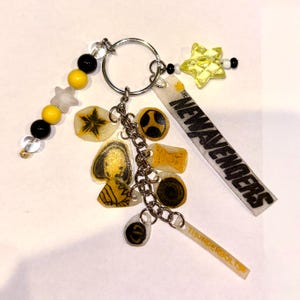 May include: A metal keychain featuring a silver ring with various charms including a yellow star, black and yellow beads, and translucent acrylic pieces. The charms display illustrations and the text THE NEWAVENGERS and THUNDERBOLTS.