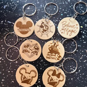 Puede incluir: Siete llaveros redondos de madera con anillas metálicas, cada uno con diseños grabados a láser que incluyen siluetas de Mickey Mouse, un timón de barco, un crucero, el texto The Stewart Family y la frase Believe In Magic.