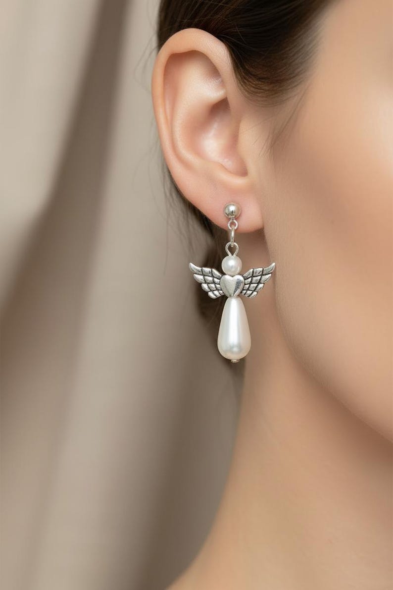 May include: Silver-toned angel earrings with pearl accents. Each earring features a small ball stud, a heart-shaped body, and detailed wings. A teardrop-shaped pearl hangs below the angel design, creating a delicate and elegant look.