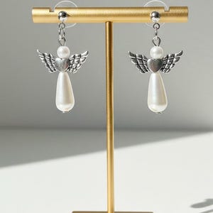 May include: A pair of angel earrings displayed on a gold stand. Each earring features a silver-toned wing design above a white pear-shaped bead. The earrings are connected with a small silver chain.