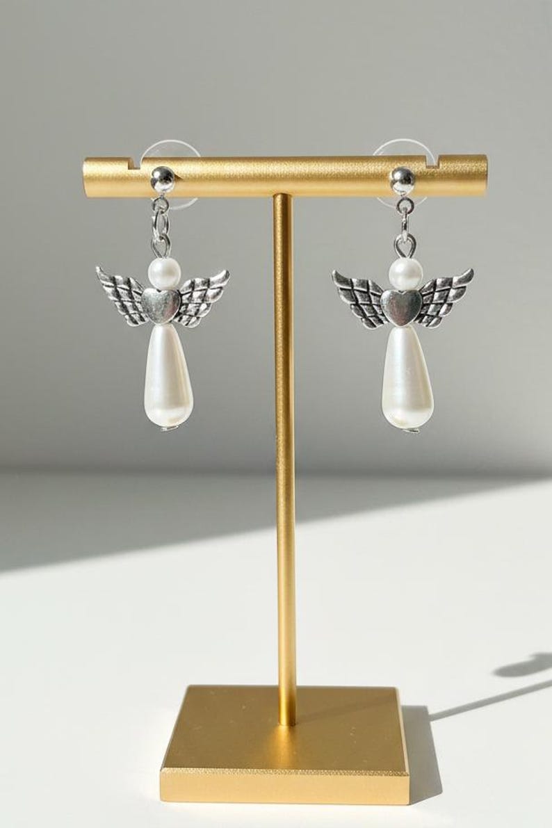 May include: A pair of angel earrings displayed on a gold stand. Each earring features a silver-toned wing design above a white pear-shaped bead. The earrings are connected with a small silver chain.