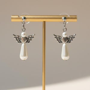 May include: A pair of silver-toned angel earrings with pearl accents. Each earring features a heart-shaped body, detailed wings, and a teardrop-shaped pearl pendant. The earrings are displayed on a gold-colored stand.