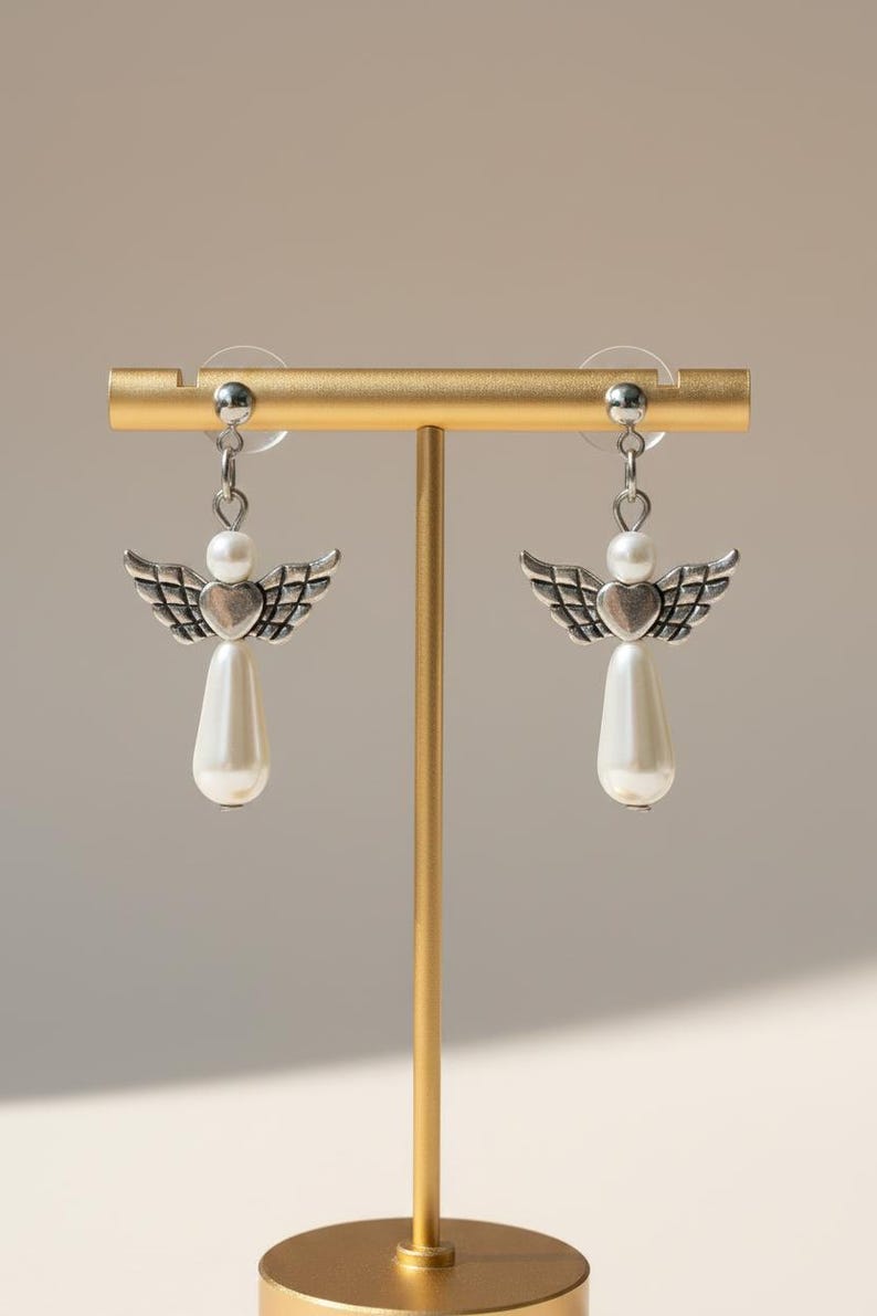 May include: A pair of silver-toned angel earrings with pearl accents. Each earring features a heart-shaped body, detailed wings, and a teardrop-shaped pearl pendant. The earrings are displayed on a gold-colored stand.